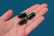 © JuliaArnhem - A hand holding two dark green supplement capsules, solid color background
