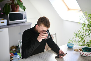 Young man sitting in the kitchen in the morning, drinking coffee and checking the news on his smartphone