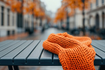 Poster - Orange knitted scarf is lying on an empty bench against the backdrop of a blurred autumn city