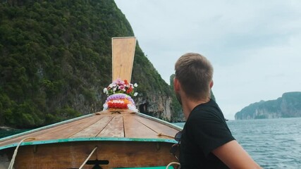 Wall Mural - Man tourist sit on bow of traditional Thai wooden long-tail boat with turquoise water and green cliffs on Phi Phi islands, Krabi, Thailand. Sailing to Maya Bay beach. Travel and tourism in Asia