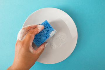 A hand holds a plate with a cleaning sponge with soap foam on a blue isolation background