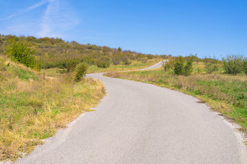 A winding rural road curves through green hills under clear blue sky during bright daylight in tranquil countryside