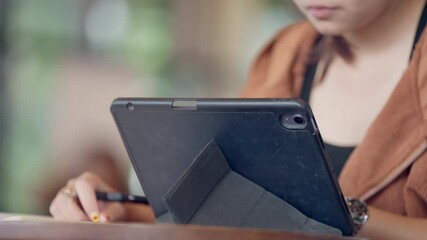 A woman is using a tablet with a black case. She is writing on a piece of paper with a pen