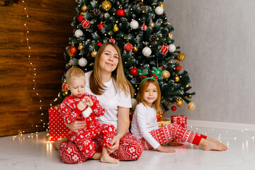 A mother and her two toddlers in festive pajamas are enjoying playtime beside the Christmas tree