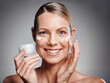 © KayExam/peopleimages.com - Portrait, happy woman and skincare cream in jar for moisturizer, beauty and collagen. Lotion, mature model and apply cosmetics on face for dermatology, glow and anti aging on gray studio background
