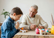 © Hanna - A young boy and his grandfather enjoy decorating Easter eggs together at a cozy table in their bright living room