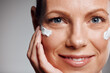 © KayExam/peopleimages.com - Portrait, mature woman and skincare cream for hydration, beauty or closeup. Lotion, happy model or hand apply cosmetics on face for dermatology, shine or glow for anti aging on gray studio background