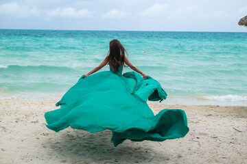 Beautiful young woman with blond long hair in green fluttering dress is standing a back on the coastline of azure caribbean sea	