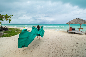 Beautiful young woman with blond long hair in green fluttering dress is standing a back on the coastline of azure caribbean sea	