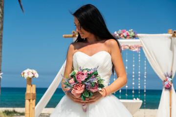 Beautiful wedding couple bride in lace wedding dress with bouquet of roses and groom reach their hands to sun near white arch on the beach of caribbean sea	