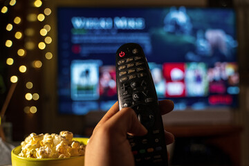 A close-up of a hand holding a TV remote control, with a TV in the background showing the online web streaming services. A bowl of popcorn is visible in the foreground.