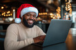 © iamfrk7 - Smiling Man Wearing Santa Hat Working on Laptop in Festive Cafe with Christmas Tree for Holiday Themed Remote Work Concept