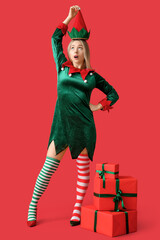 Wall Mural - Beautiful young woman in elf costume with Christmas gift boxes on red background