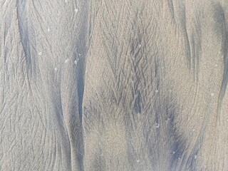 Natural patterns in the beach sand created by wind and waves, highlighted by soft, warm sunlight, form intricate lines and textures in a serene coastal setting.