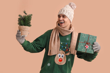 Poster - Young man in Christmas sweater with fir tree and gift on beige background