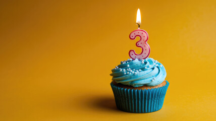 A blue cupcake with a lit candle on top that says 3. The cupcake is on a yellow background