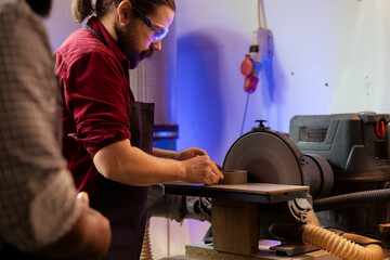 Carpenter in woodworking shop wearing protection glasses while shaping wooden object on disc sander. Cabinetmaker with safety equipment in joinery using professional grinding machine