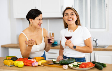 Happy female friends with glasses of red wine chatting and preparing salad together in modern kitchen