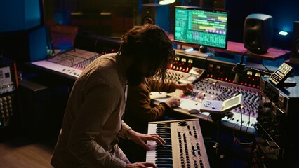 Poster - Skilled musician performing in control room at studio and recording tracks for new album, working with audio console and flying faders. Songwriter composing new music on technical tools. Camera B.