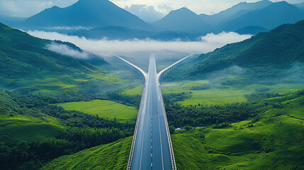 Aerial view of a highway bridge in a green valley, with a mountain range in the background and a foggy sky