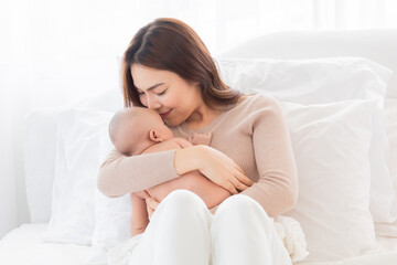 Asian young mother holding tenderly cuddles newborn baby in arms on to chest. Mother looking adorable infant with love and care. Woman and small child lying on bed together at home