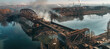 © Anoo - Aerial view of the damaged bridge over the river with smoke rising from an explosion against the city skyline in daylight