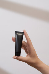 Sticker - Woman's hand holds a mockup of black tube of cream. Unlabeled packaging for cosmetics in sunlight on white background. Concept of skincare. Harsh shadows