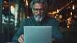 ©  Jovaduplex - Mature man with glasses deeply focused on his laptop work in an intimate, warmly lit cafe environment, suggesting a cozy and relaxed atmosphere.