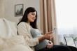© Lena Ivanova - Beautiful smiling pregnant woman with baby belly use mobile cell phone, do online shopping, browse internet, watch podcast while sitting on sofa at home. Having doctor video call medical appointment