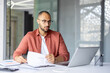 © Liubomir - Confident businessman working on laptop, analyzing documents and focusing on important data at office desk. Professional setting with modern design elements.