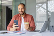 © Liubomir - Smiling man holding smartphone and credit card while online shopping at office desk. Engaged in digital purchasing with phone, displaying convenience and modern technology.