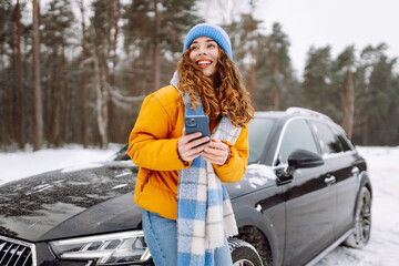 young woman with a phone near her car in a winter forest. sending messages, communicating with frien