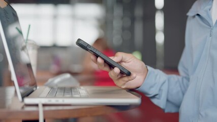 A man is holding a cell phone while sitting at a table with a laptop. The man is looking at the phone, possibly checking a message or browsing the internet. The scene suggests a moment of multitasking