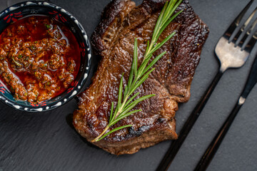 Grilled Marbled Beef Steak with a sprig of rosemary on a mica board with sauce and Argentinian Chimichurri sauce in a bowl on a dark background