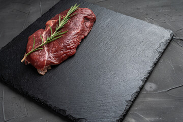 Raw organic marbled beef steak with a sprig of rasmarin on a black mica board on a dark background. Copyspace.