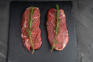 Two pieces of raw meat for steaks with a sprig of rosemary on a black mica board on a dark background.