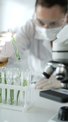 Woman scientist with white gloves, face mask and protective glasses, is holding a test tube with plants inside near microscope in laboratory setting, vertical portrait view. Science and medicine