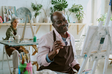 Wall Mural - African American jolly man sitting in front of wooden easel with canvas and enjoying painting in drawing studio