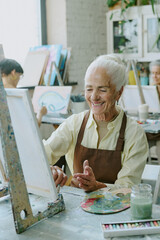 Wall Mural - Vertical shot of silver haired woman smiling widely while doing strokes of paint brush on white canvas , she sitting at retro table with drawing tools