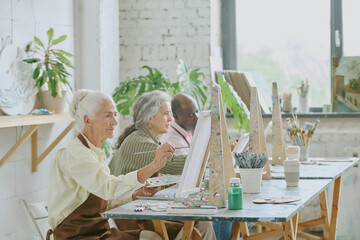 Wall Mural - Senior biracial people sitting at dirty wooden table with easel, canvas and painting tools while painting together