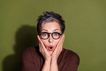 Wall Mural - Photo portrait of gray hair beautiful cheerful pensioner grandmother in brown t shirt touching cheeks amazed isolated on khaki color background