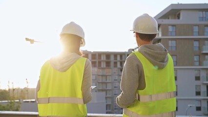 Two engineers are standing on a construction site wearing hard hats and safety vests looking at a building and discussing something