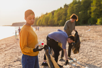 Earth day. Volunteers activists team collects garbage cleaning of beach coastal zone. Woman puts plastic bottle trash in garbage bag on ocean shore. Environmental conservation coastal zone cleaning