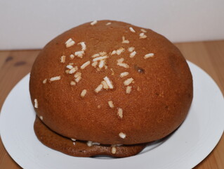 A large brown cake with white sprinkles on top sits on a white plate. The cake is round and he is a dessert