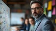 © cynz - A man in his early 40s, wearing a gray suit and glasses, stands at a whiteboard, his face filled with determination as he leads a critical brainstorming session. His colleagues, a mix of men and