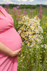 A expectant mother cradles her round belly while holding a vibrant bouquet of daisies amidst a blooming wildflower field under a bright blue sky. 