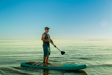 A man in sports shorts and a T-shirt on a SUP board with a paddle in a calm sea against the background