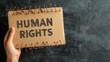 © adynue - hand holding cardboard paper with HUMAN RIGHTS text and rusty sharp bare wire on dark background, human rights day concept