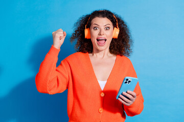 Wall Mural - Photo of wavy haired model woman wearing orange cardigan fist up listen headphones wireless connection isolated on blue color background