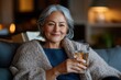 © Milos - A confident mature woman sits relaxed in a cozy living room, enjoying a refreshing drink, wrapped in a warm sweater in the inviting comfort of her home.